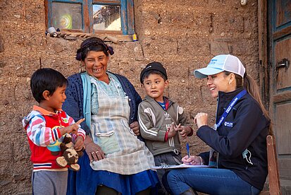 Eine Frau sitzt vor einer Lehmwand neben zwei Kindern, gegenüber sitzt eine Mitarbeiterin von Plan mit Kappe und Notizblock auf einem Stuhl; alle schauen einander an, lachen und gestikulieren.