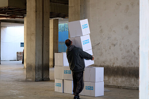 Ein Mann stapelt mehrere Kisten mit Hilfsgütern von Plan International in einer Halle übereinander