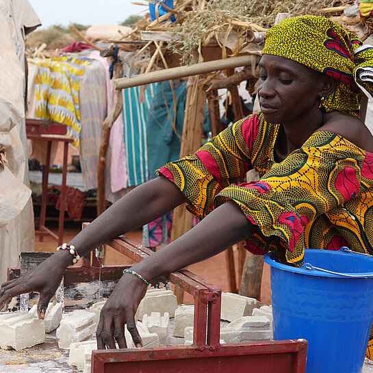 Eine Frau (Hamssetou) steht an einer Werkbank und breitet Seifenstücke aus, um sie in verkaufsfertige Portionen zu zerschneiden.