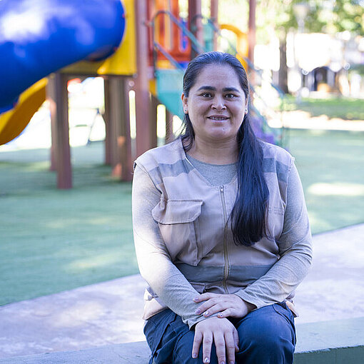 Porträt von Rosana auf einem Spielplatz. Sie steht im Freien vor bunten Spielgeräten und blickt in die Kamera.