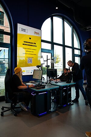 Szene in einer modernen Halle mit hohen Fenstern. Links sitzt eine Person am Computer, rechts stehen zwei Personen - einer spricht ins Mikrofon. Im Hintergrund ein großes gelbes Plakat: „Gemeinsam für Menschenrechte! Dieser Stream sammelt Spenden für Amnesty International.“ QR-Code ist sichtbar, mit Bisalina Speedruns Logo und Sponsorenlogos.