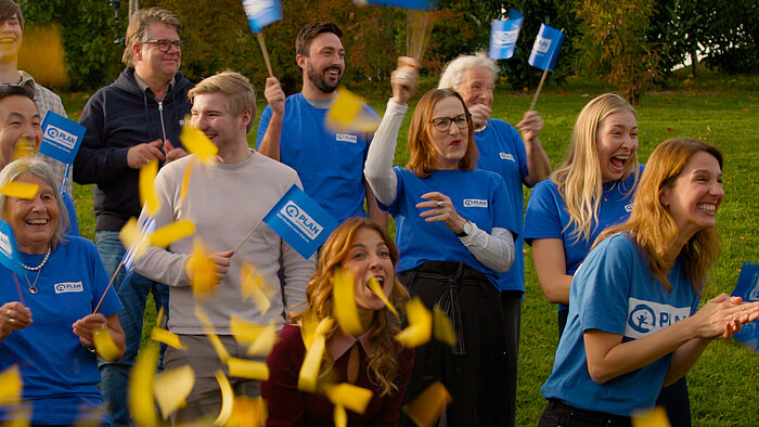 Eine Gruppe von Menschen in blauen Plan-T-Shirts steht auf einer Wiese und jubelt. Sie halten kleine Plan-Flaggen, während gelbe Konfetti in die Luft fliegen. Die Stimmung ist fröhlich und feierlich.