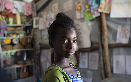 Ein Mädchen schaut ernst in die Kamera. Hinter hier hängen bunte Bilder an der Wand.