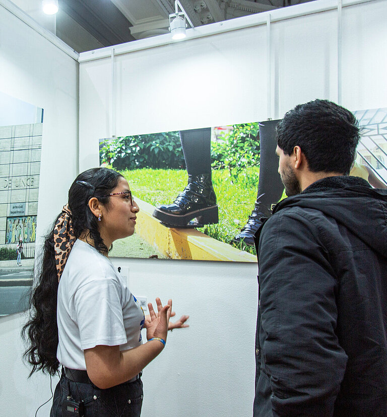 Eine junge Frau steht vor einem Foto, das in Großformat an einer Wand hängt, und spricht mit einem Mann, der neben ihr steht