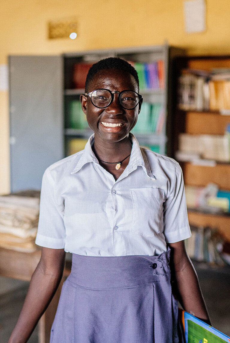 Moreen, eine junge Frau in Schuluniform und Brille, steht strahlend in der Schulbibliothek.
