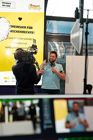  Szene beim Livestream. Ein Moderator mit Mikrofon steht vor einer Kamera und spricht in den Raum. Im Hintergrund ein gelbes Plakat mit „Gemeinsam für Menschenrechte!“, Bisalina Speedruns Logo, Spenden-QR-Code und Sponsorenlogos. Im Vordergrund sind Kameras und technische Geräte.