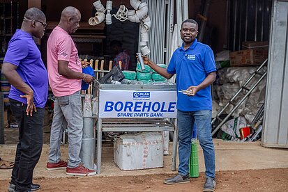 Plan-Mitarbeiter stehen vor einem Ersatzteillager für Wasseranlagen mit dem Schild „Borehole Spare Parts Bank“ – ein Projekt zur nachhaltigen Wartung von Brunnen.