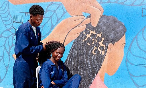 Ein junger Auszubildender arbeitet draußen an der Flechtfrisur einer jungen Frau