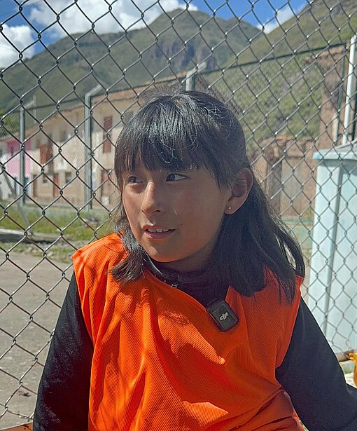 Ein Mädchen mit orangefarbener Weste steht an einem Sportplatz. 