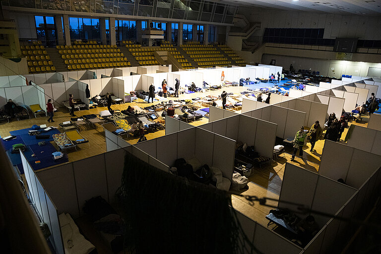 Eine Sporthalle wurde durch Stehwände in einzelne Schlafplätze unterteilt. Auf dem Boden liegen Matratzen, einige Menschen laufen durch den Raum.