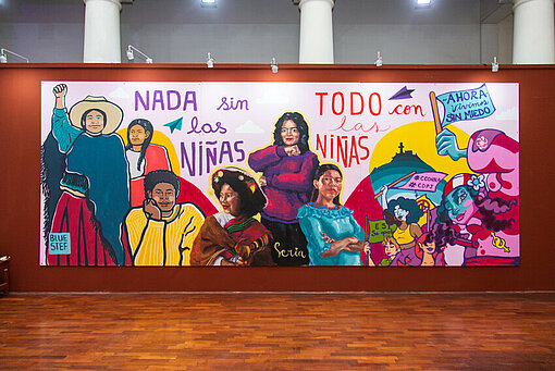 Wandgemälde im Kulturzentrum für Bildende Künste in Lima zum Weltmädchentag 2022. Das Thema: „Wir sind hier: Die Stimme von Mädchen für politische Teilhabe“ Ein Wandgemälde in einer Kunstgalerie, auf dem die Geschichte der Mädchenrechte in Peru graphisch dargestellt ist. Frauen in kämpferischen Posen und Schriftzüge sind zu sehen.