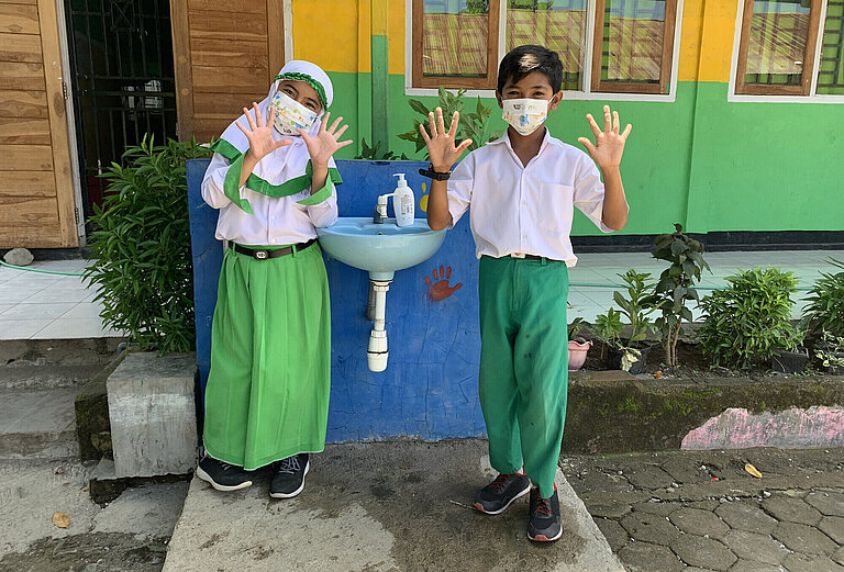 Nadia und Ceni stehen neben einem Waschbecken und winken in die Kamera. Sie tragen medizinische Masken und ihre Schuluniform.