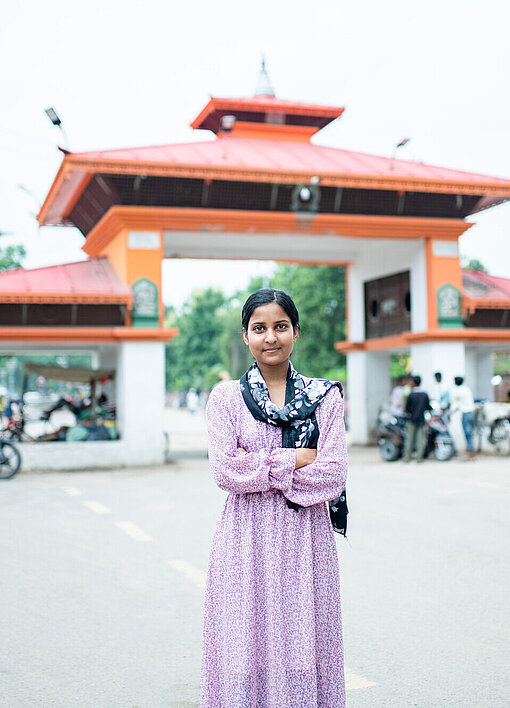 Eine junge Frau steht auf der Straße vor einem großen Tor. Im Hintergrund sind Menschen und Mopeds