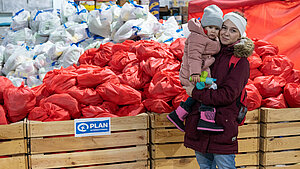 Mutter mit Kind vor einem großen Haufen von Hilfspaketen in einem Lagerraum von Plan International in der Ukraine