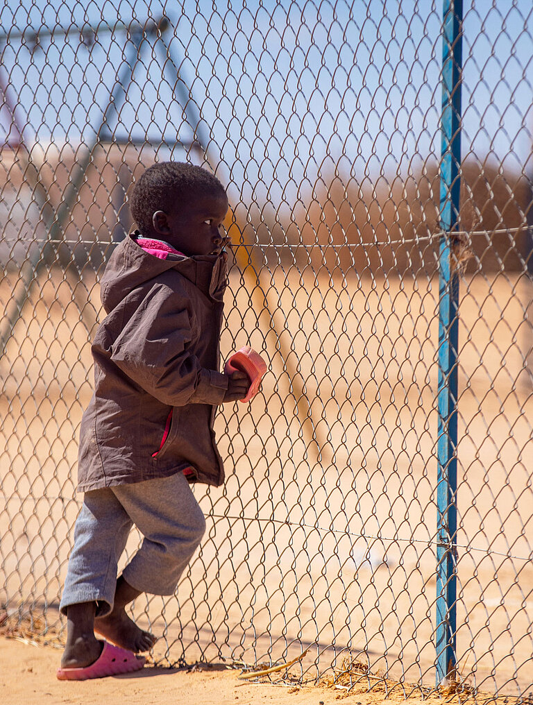 Ein kleines Kind hält sich mit einer Hand an einem Maschendrahtzaun fest und schaut hindurch.