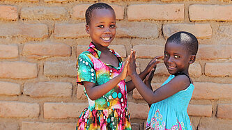 Zwei Mädchen stehen sich gegenüber und halten ihre Hände aneinander. Sie lächeln in die Kamera.