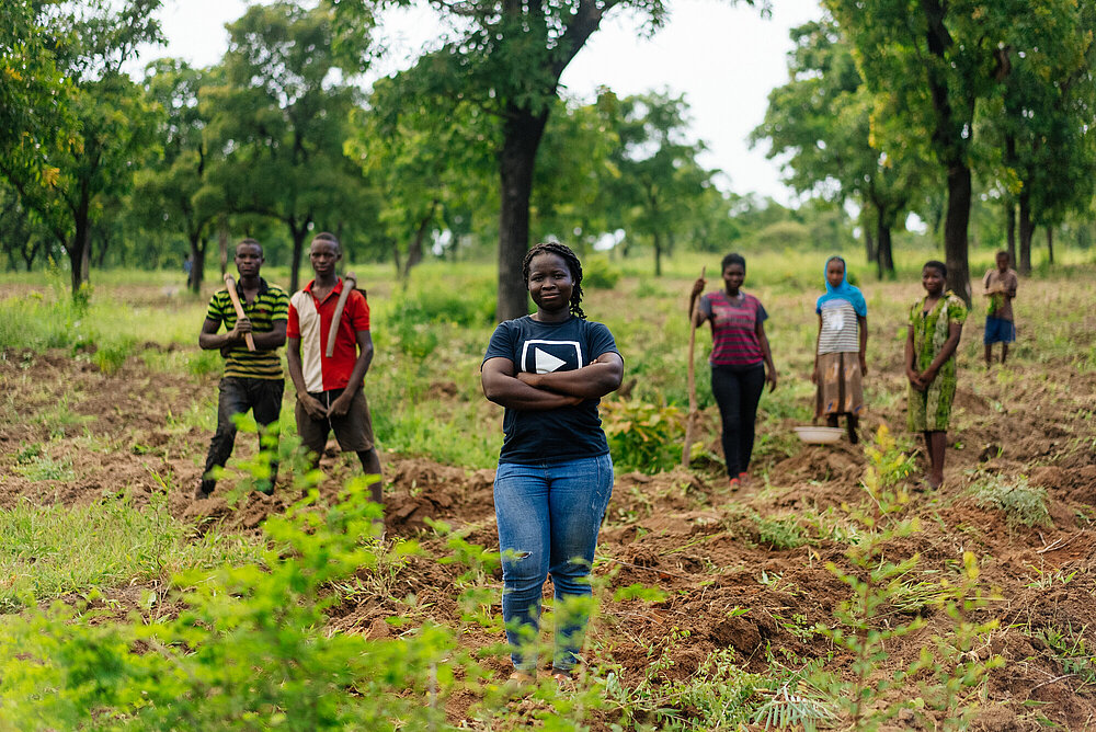 Junge Frau steht selbstbewusst vor einer Gruppe Jugendlicher, die auf einem Feld in Ghana landwirtschaftlich arbeiten