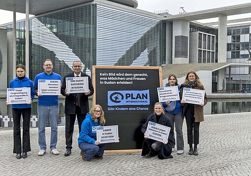 Mehrere Personen mit blauen Jacken und dem Plan-Logo haben sich am Ufer der Spree vor einem Bürogebäude versammelt.