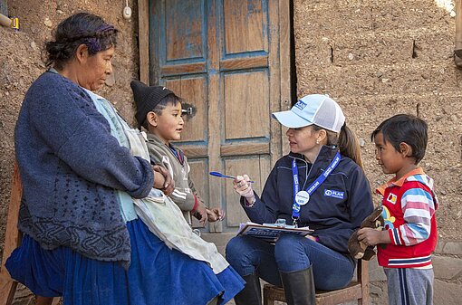 Eine Frau mit Plan-Logo an der Bekleidung befragt eine Familie und notiert die Antworten in einer Projektdokumentation