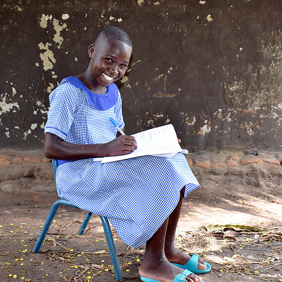 Mädchen in Schuluniform sitzt draußen und macht Hausaufgaben, lächelt in die Kamera