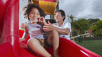 Ein Mann steht an einer roten Plastikrutsche und behütet seine rutschende Tochter mit lockigen Haaren