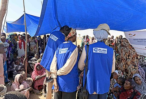 Zwei Frauen und ein Mann mit blauen Westen mit dem Plan-Logo drauf stehen unter einem blauen Zeltdach und sprechen mit Dutzenden Frauen.