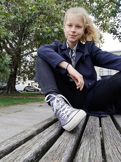 Eine junge Frau sitzt mit angewinkeltem Bein auf einer Bank und schaut in die Kamera