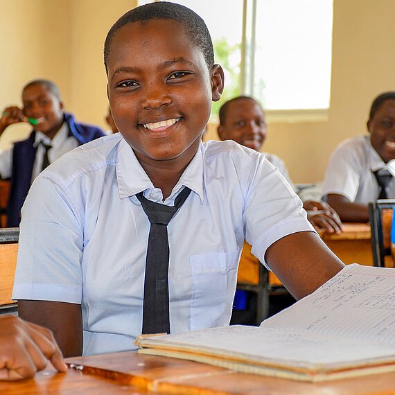 Ein 16-jähriges Mädchen aus Tansania sitzt mit weiteren Kindern in ihrem Klassenraum vor aufgeschlagenem Buch. Sie trägt ihre Schuluniform und hat ein breites Lächeln im Gesicht.