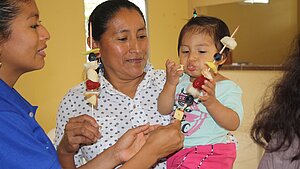 Eine Frau hält ein Kleinkind im Arm, das fröhlich einen Fruchtspieß isst. Weitere Erwachsene reichen Obststücke – Szene aus einem Gesundheitsprojekt in Ecuador.