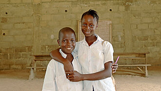 In einem kahlen Raum stehen ein Mädchen und ein Junge vor einer improvisierten Schultafel