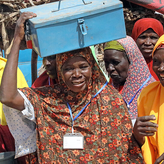Eine Gruppe von Frauen in bunten Gewändern hat sich auf einem Dorfplatz versammelt. Eine Frau in der Mitte der Gruppe trägt eine metallene Geldkassette auf dem Kopf.