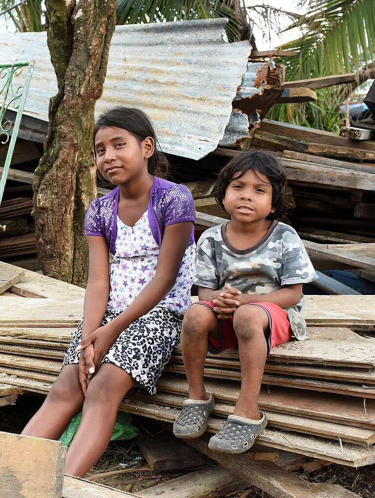 Zwei Kinder sitzen auf Holzbalken vor einem kaputten Haus.