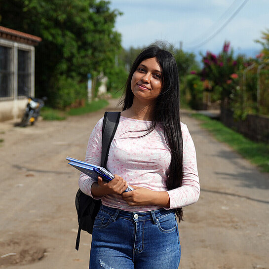 Teenagerin trägt Schulrucksack und Notizblock, unterwegs in einer ländlichen Gegend. Bildung als Schlüssel für die Zukunft.
