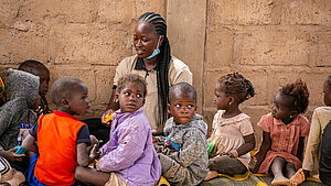  Lehrerin sitzt mit Kleinkindern auf Matten im Kreis und unterrichtet sie spielerisch – Vorschule in Westafrika