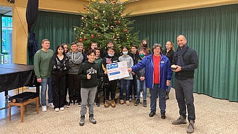 Die Spenden wurden an Marlies Grundmann von der Plan-Aktionsgruppe Saarbrücken übergeben. Bild: Eine Gruppe von Menschen steht vor einem Weihnachtsbaum und halten einen Plan-Scheck in die Höhe.