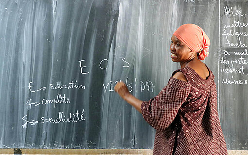 Eine Frau in rot-braunem Kleid schreibt an einer Schultafel