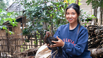 Eine 23-Jährige aus Laos steht draußen und hält ein Huhn in den Händen, hinter ihr sind Holzhütten und grüne Pflanzen
