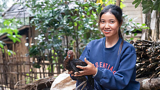 Eine 23-Jährige aus Laos steht draußen und hält ein Huhn in den Händen, hinter ihr sind Holzhütten und grüne Pflanzen