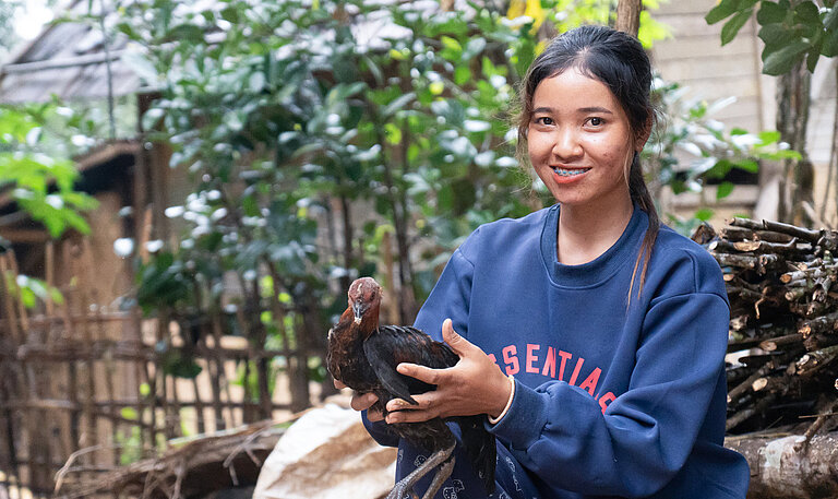 Eine 23-Jährige aus Laos steht draußen und hält ein Huhn in den Händen, hinter ihr sind Holzhütten und grüne Pflanzen