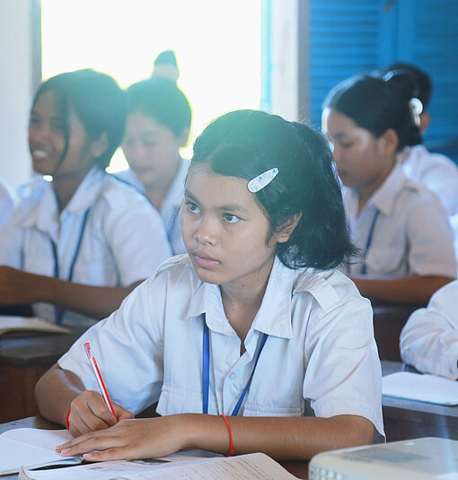 Schülerinnen in weißen Hemden sitzen im Klassenraum und schreiben in ihre Hefte, ein Mädchen ist im Fokus