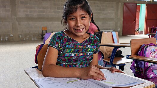 Lächelndes Mädchen sitzt in einem Klassenzimmer mit Heften auf ihrem Tisch
