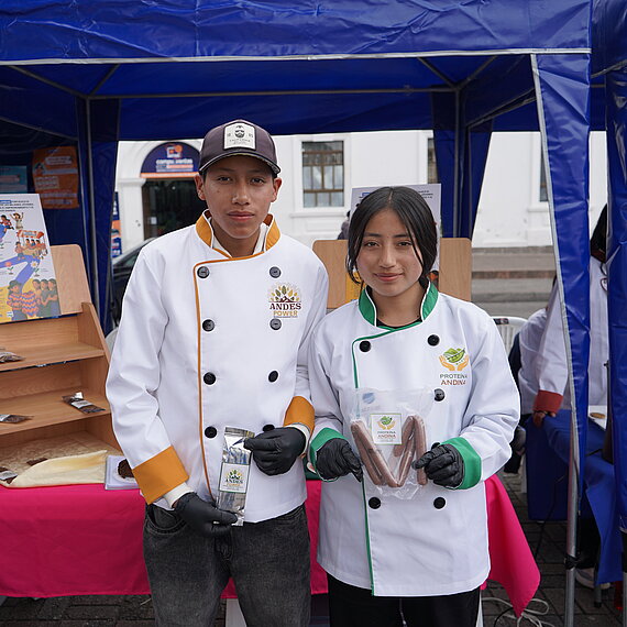 Ein junger Mann und eine junge Frau, beide gekleidet in professionelle weiße Kochjacken, stehen nebeneinander und halten jeweils Lebensmittelverpackungen in den Händen. Sie wirken stolz und selbstbewusst, als würden sie ihre Produkte auf einer Messe oder Ausstellung präsentieren