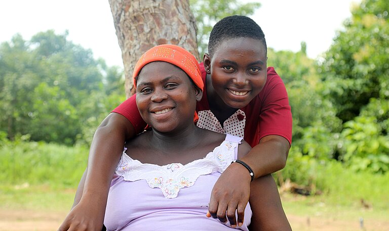Eine junge Frau mit rotem T-Shirt umarmt eine ältere Frau, die im Garten auf einer Bank sitzt und ein weißes Shirt trägt, und beide lächeln