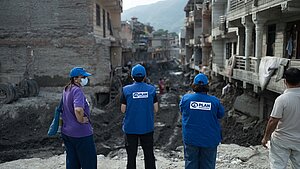 Drei Personen mit blauen Kappen und blauen Westen mit dem Logo von Plan International sowie eine weitere Person stehen vor einer zerstörten, schlammigen Straße zwischen beschädigten Häusern. Im Hintergrund sind weitere Menschen und Berge zu sehen.