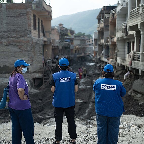 Drei Personen mit blauen Kappen und blauen Westen mit dem Logo von Plan International sowie eine weitere Person stehen vor einer zerstörten, schlammigen Straße zwischen beschädigten Häusern. Im Hintergrund sind weitere Menschen und Berge zu sehen.
