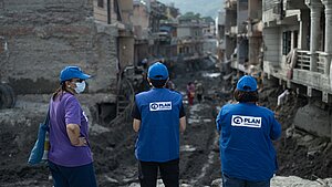 Drei Personen mit blauen Kappen und blauen Westen mit dem Logo von Plan International sowie eine weitere Person stehen vor einer zerstörten, schlammigen Straße zwischen beschädigten Häusern. Im Hintergrund sind weitere Menschen und Berge zu sehen.