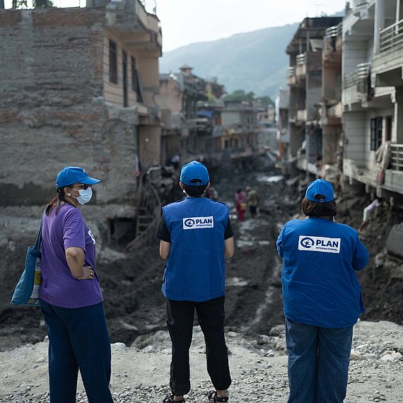 Drei Personen mit blauen Kappen und blauen Westen mit dem Logo von Plan International sowie eine weitere Person stehen vor einer zerstörten, schlammigen Straße zwischen beschädigten Häusern. Im Hintergrund sind weitere Menschen und Berge zu sehen.