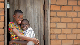 Zwei lachende Kinder stehen eng umarmt vor einer einfachen Ziegelwand und einer Holztür und schauen fröhlich in die Kamera.