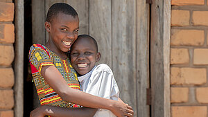 Zwei lachende Kinder stehen eng umarmt vor einer einfachen Ziegelwand und einer Holztür und schauen fröhlich in die Kamera.