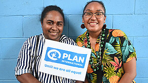 Zwei Frauen auf den Salomonen lächeln in die Kamera und halten ein Schild mit dem Plan-International-Logo und dem Slogan ‚Until we are all equal‘.